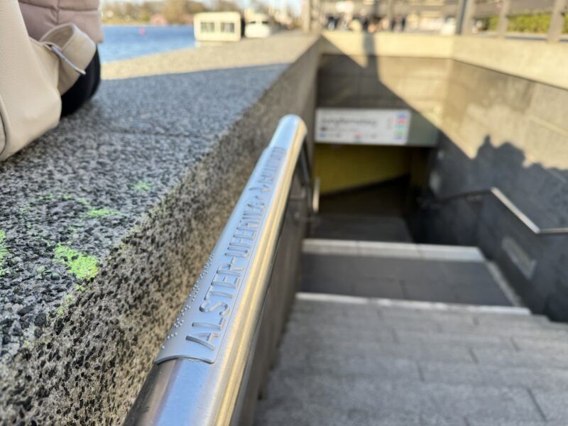 Foto eines Handlaufs, der mit einer Plakette ausgestattet wurde. In Klar- und Braillie-Schrift markiert sie den Standort Alster-Uferweg. Im Hintergrund die Binnenalster.