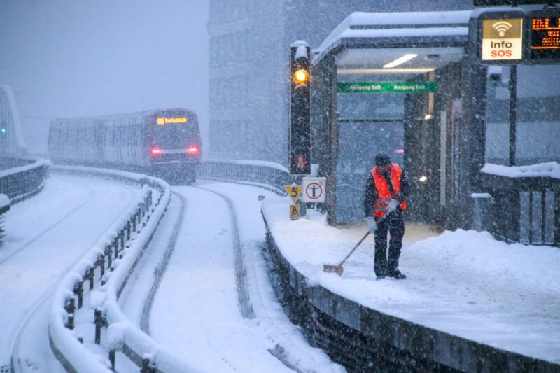 Foto eines einfahrenden U3-Zuges. Auf dem Bahnsteig ein Kollege beim Schneeräumen.